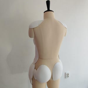May include: A beige dress form with a black neck piece and white padding. The dress form is on a metal stand. The white padding is in the shape of shoulder pads and other body shaping pieces.