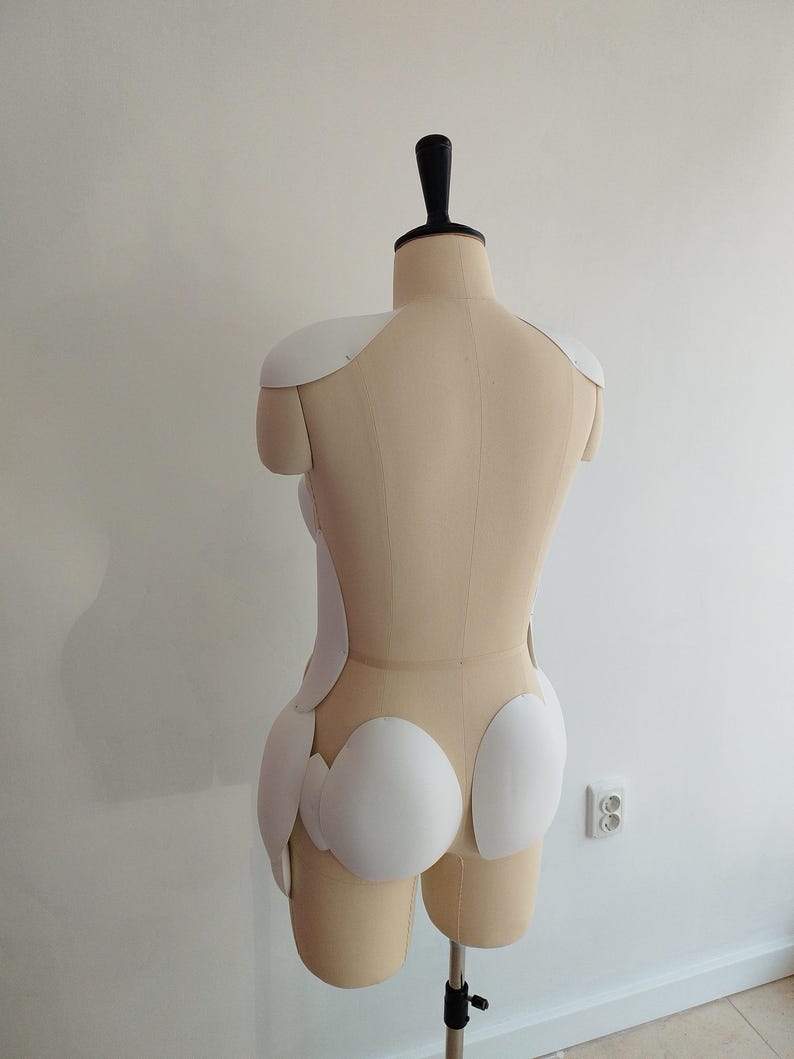 May include: A beige dress form with a black neck piece and white padding. The dress form is on a metal stand. The white padding is in the shape of shoulder pads and other body shaping pieces.