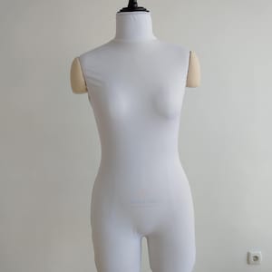 Puede incluir: Un maniquí blanco con cuello y soporte negros. El maniquí tiene un torso redondeado, hombreras y cuello. El soporte es de metal. Se utiliza para exhibir ropa.