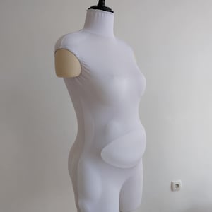 Op de afbeelding: Een witte paspop met een zwarte nek en standaard. De vorm is bedekt met witte stof en heeft een gewatteerd gedeelte voor de buik. De vorm is ontworpen om een zwanger lichaam te simuleren.