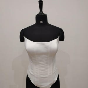 May include: A white satin bodysuit displayed on a black dress form. The bodysuit has a curved neckline and a fitted bodice. The dress form has the number 86 on the neck.