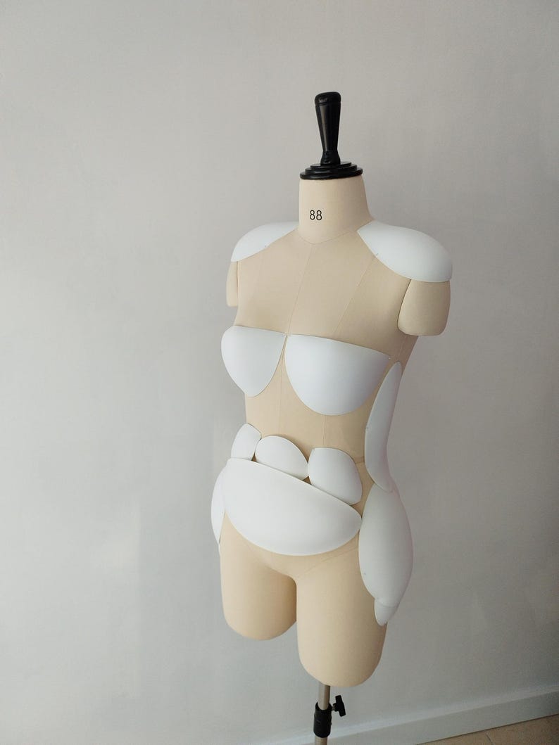 May include: A beige dress form with white padding and a black neck piece. The dress form has the number 88 printed on the neck. The form is on a black stand, and the background is a plain white wall.
