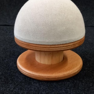 May include: A mushroom-shaped pin cushion with a light gray fabric top and a wooden base. The base has a round, tiered design with a natural wood finish. The pin cushion is designed for sewing and crafting.