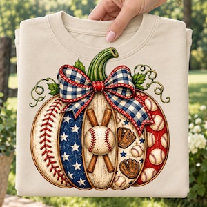 Op de afbeelding: Een beige t-shirt met ronde hals en een afbeelding van een pompoen versierd met honkbal-elementen. De pompoen heeft secties met honkbalstiksels, blauwe sterren, houtnerf en rode patronen met honkballen en handschoenen, afgewerkt met een strik.