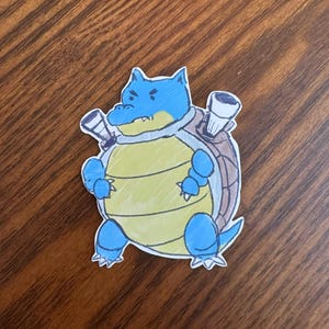 Puede incluir: Una pegatina troquelada con una ilustración dibujada a mano de una criatura bípeda azul y amarilla con un caparazón marrón en la espalda. El personaje tiene dos grandes cañones que sobresalen del caparazón, una barriga amarilla clara y extremidades azules.
