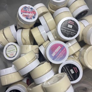 Puede incluir: Una colección de pequeños frascos redondos llenos de una sustancia cremosa de color amarillo pálido. Los frascos tienen tapas blancas con etiquetas con textos como "NOIR" y "Baby Powder Whipped Body Butter". Algunas etiquetas muestran diseños florales.