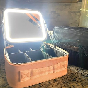 Puede incluir: Un estuche de maquillaje de color beige con un espejo y una luz incorporados. El estuche tiene una cremallera y un asa. El estuche está abierto y el interior es visible, mostrando compartimentos para guardar maquillaje.