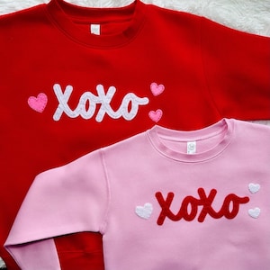 May include: Two sweatshirts, one red and one pink, with the word "XOXO" in white felt on the red sweatshirt and red felt on the pink sweatshirt. Both sweatshirts have small pink felt hearts.