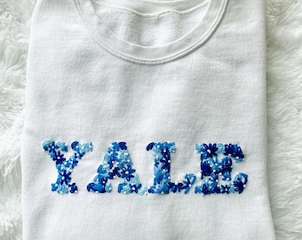 May include: White sweatshirt with the word "YALE" embroidered in blue and white flowers.