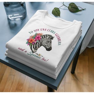 May include: A stack of white t-shirts featuring a zebra head with floral accents and the text "Yo soy una cebra guerrera and a Spoonie too!" The zebra has black and white stripes and is surrounded by pink, orange, and red flowers.