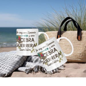 May include: Two white ceramic mugs with the text "Jo soy una Spoonie y una CEBRA GUERRERA también" are displayed on a beach. The mugs have a floral and zebra design. A woven tote bag with black handles and a gray blanket are also in the image.