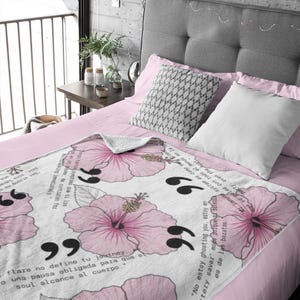 May include: A white blanket with a repeating pattern of large pink hibiscus flowers and black quotation marks. The blanket is draped over a bed with pink sheets and pillows. Text is printed on the blanket.