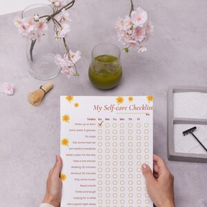 May include: A self-care checklist with floral accents is held by a person. The checklist includes tasks like drinking water, doing yoga, and getting fresh air. A glass of green liquid and a branch with pink flowers are also visible.