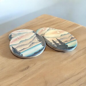 May include: Set of four round wooden coasters with a painted design of a mountain range, a lake, and trees at sunset. The coasters have a light wood background with a light blue, pink, and orange sky.