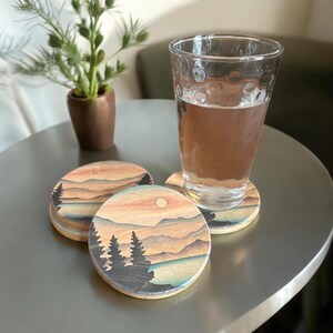 May include: Set of three round coasters with a sunset landscape design. The coasters feature a mountain range, a lake, and a sun setting behind the mountains. The coasters are made of a light brown material and have a slightly textured surface.