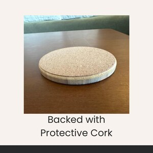 May include: A round coaster with a cork backing and a light-colored wooden edge. The coaster is on a brown wooden surface. The text "Backed with Protective Cork" is below the coaster.