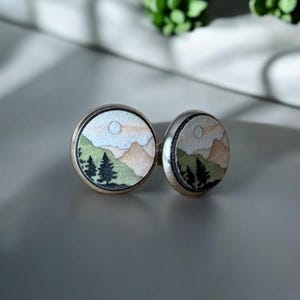 May include: A pair of round, silver cufflinks with a painted landscape scene. The scene features a mountain range with a sun and clouds in the sky, and a group of trees in the foreground. The cufflinks are a light silver color.