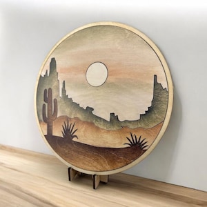 May include: A round wooden wall hanging with a desert scene. The scene features a large, full moon, a cactus, and a mountain range in the background. The wood is stained in shades of brown, green, and orange.