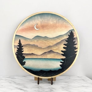 May include: Round wooden wall art featuring a landscape scene. The artwork depicts a lake, mountains, and trees under a sunset sky with a crescent moon. The piece has a natural wood frame and is displayed on a wooden stand.