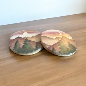 May include: Two wooden coasters with a painted landscape scene. The design features a sunset with mountains, trees, and a moon. The coasters are oval-shaped and made of light-colored wood.
