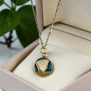 May include: A gold-colored necklace with a circular pendant. The pendant features a landscape design with a white mountain, blue sky, and yellow and green foreground. The necklace is displayed in a pink jewelry box.