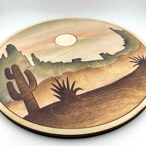 May include: A round wooden wall hanging with a desert scene. The scene features a cactus, a yucca plant, and a mountain range in the background. The sun is setting behind the mountains, casting a warm glow on the scene.