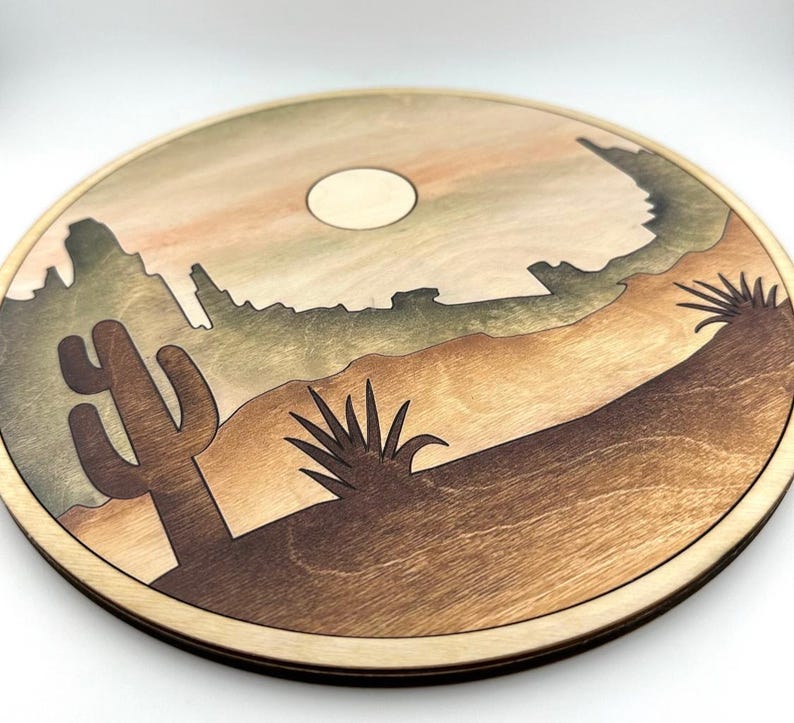 May include: A round wooden wall hanging with a desert scene. The scene features a cactus, a yucca plant, and a mountain range in the background. The sun is setting behind the mountains, casting a warm glow on the scene.