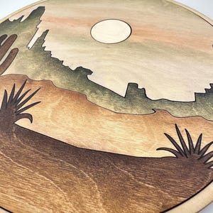 May include: A round wooden wall art piece with a desert landscape scene. The scene features a mountain range, a full moon, and two desert plants. The colors are brown, green, and beige.