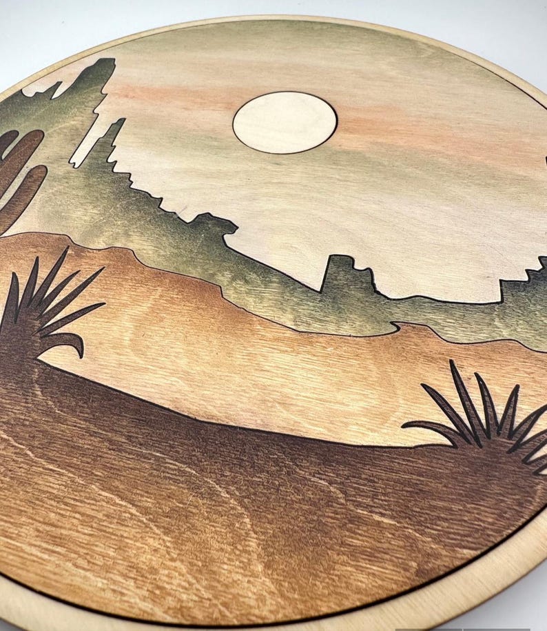 May include: A round wooden wall art piece with a desert landscape scene. The scene features a mountain range, a full moon, and two desert plants. The colors are brown, green, and beige.