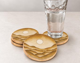 May include: Three round wooden coasters featuring a laser-engraved landscape design of rolling hills and a sun, with one clear glass tumbler filled with water resting on top of the stack of coasters, showcasing a natural light wood grain finish.