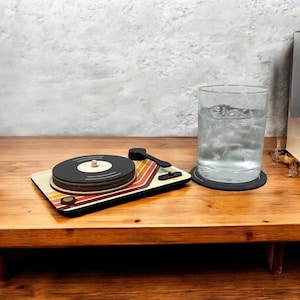 May include: A wooden turntable-shaped coaster set with a retro design. The coaster features a black record design with a beige center and a striped pattern in red, orange, and beige. A glass of water sits nearby.