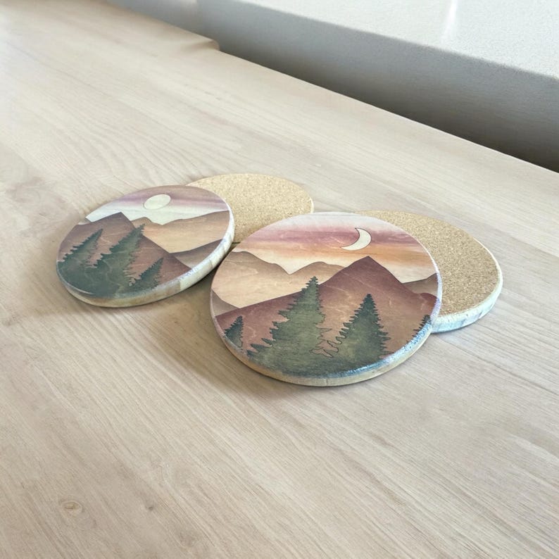 May include: Four round coasters with a mountain and forest design. The coasters feature a watercolor painting of a mountain range, trees, and a moon or sun. The color palette includes shades of brown, green, and orange.