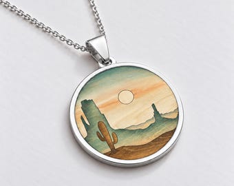 May include: A silver-toned circular pendant necklace featuring a desert landscape illustration on a wood-grain background. The design depicts a saguaro cactus, rock formations, and a sun in shades of orange, blue, and brown.