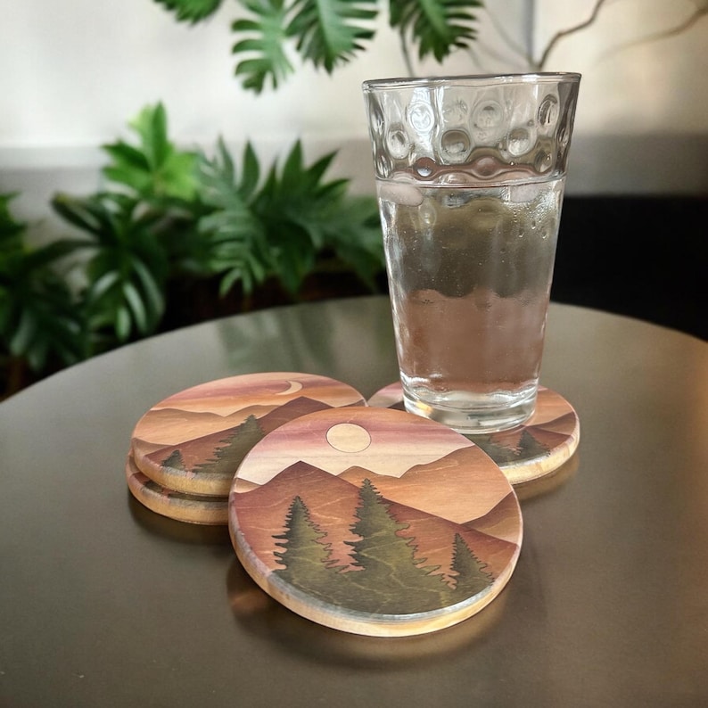 May include: Set of three wooden coasters with a landscape design featuring mountains, trees, and a moon or sun. The coasters are round and have a natural wood grain edge. A glass of water sits on one of the coasters.