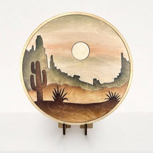 May include: A round wooden wall hanging with a desert scene. The scene features a silhouette of mountains, a cactus, and a yucca plant. The background is a gradient of brown, orange, and green.