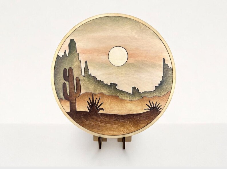 May include: A round wooden wall hanging with a desert scene. The scene features a silhouette of mountains, a cactus, and a yucca plant. The background is a gradient of brown, orange, and green.