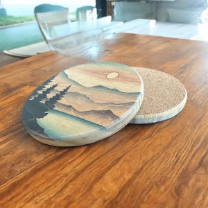 May include: Two round coasters, one with a painted design of a mountain range with a sunset and the other with a cork surface.