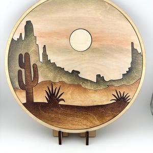 May include: A round wooden wall art piece with a desert scene. The scene features a cactus, a mountain range, and a full moon in a light brown sky. The wood is stained in shades of brown and green.