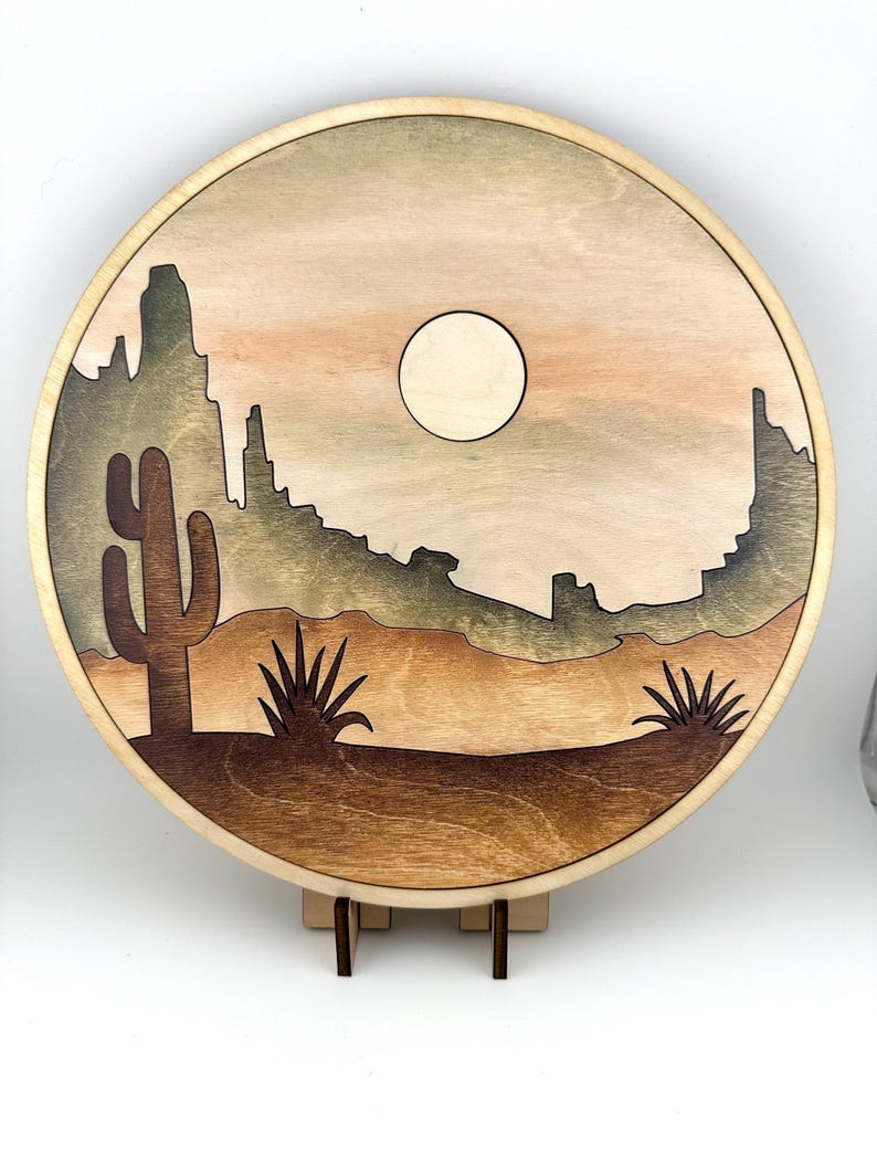 May include: A round wooden wall art piece with a desert scene. The scene features a cactus, a mountain range, and a full moon in a light brown sky. The wood is stained in shades of brown and green.