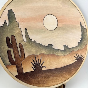 May include: A wooden wall hanging with a desert scene. The scene features a large cactus, two smaller plants, a mountain range, and a full moon in a light brown sky.