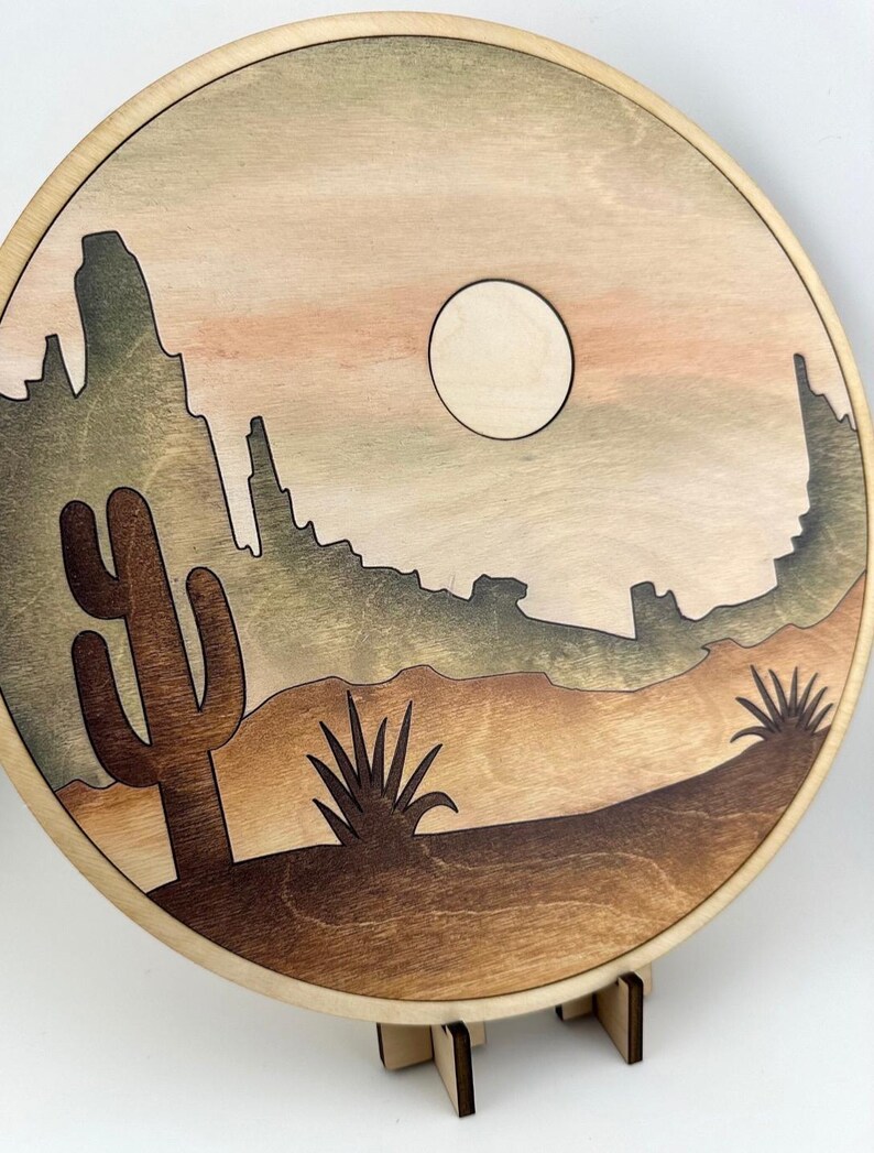 May include: A wooden wall hanging with a desert scene. The scene features a large cactus, two smaller plants, a mountain range, and a full moon in a light brown sky.