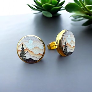 May include: A pair of gold-toned earrings with a round, white, and brown landscape design. The landscape features a mountain range, a single tree, and a white moon.