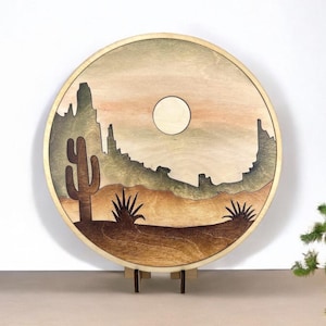 May include: A round wooden wall art piece with a desert landscape scene. The art features a silhouette of mountains, a cactus, and a moon in a light brown, tan, and green color scheme.