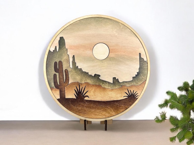 May include: A round wooden wall art piece with a desert landscape scene. The art features a silhouette of mountains, a cactus, and a moon in a light brown, tan, and green color scheme.