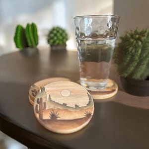 May include: Wooden coasters with a desert landscape design. The coasters feature a cactus, mountains, and a sun. A glass of water sits on one coaster. The coasters are round and made of light-colored wood.
