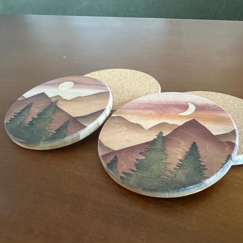 May include: Two wooden coasters with a mountain and forest landscape design. One coaster is oval, the other is round. The artwork features a sunset with a crescent moon and evergreen trees. The coasters are on a brown surface.