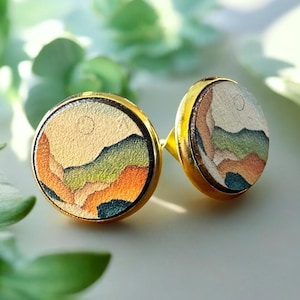 May include: A pair of gold-toned stud earrings with a round, painted design. The design features a landscape with a sun, mountains, and a sky in shades of orange, green, and blue.