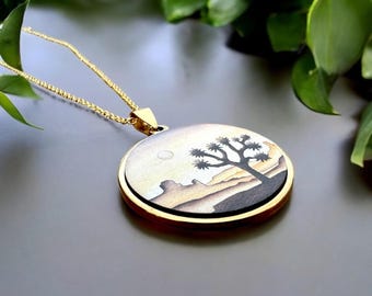 May include: A gold necklace with a round pendant featuring a hand-painted illustration of a Joshua Tree in a desert landscape. The pendant has a gold rim and a black background.