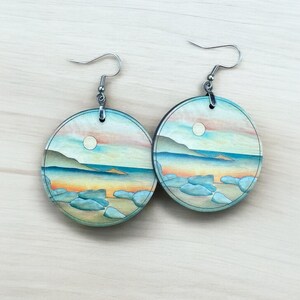 May include: Round earrings with a beach scene design. Each earring features a painted landscape with a blue ocean, rocks, and a sunset sky. The earrings have silver-colored ear wires.