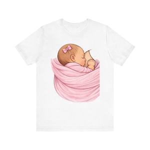 May include: White t-shirt featuring a watercolor illustration of a baby wrapped in a pink blanket, nursing. The baby has a pink bow in their hair. The design is centered on the shirt.